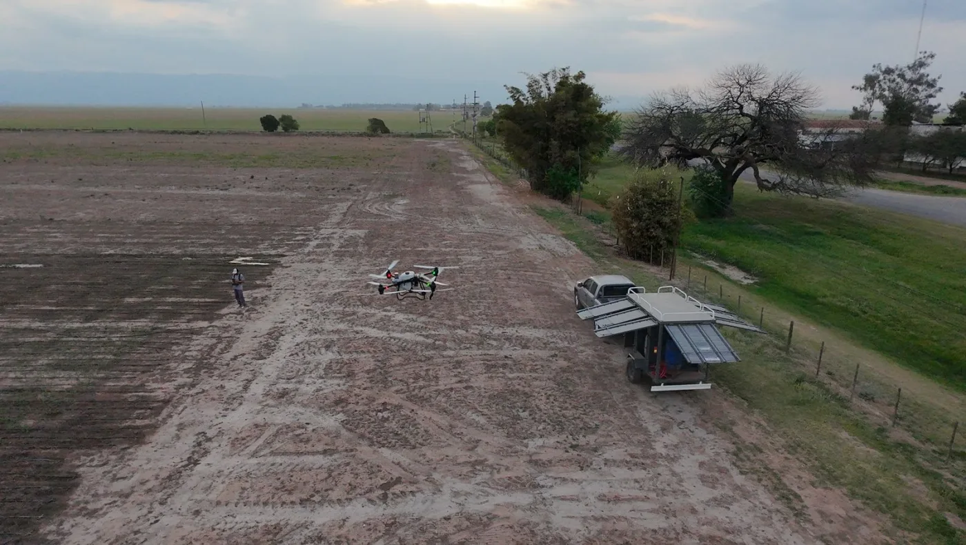 Vista aérea de un drone de pulverización en vuelo sobre un lote, con el operario y el tráiler de apoyo